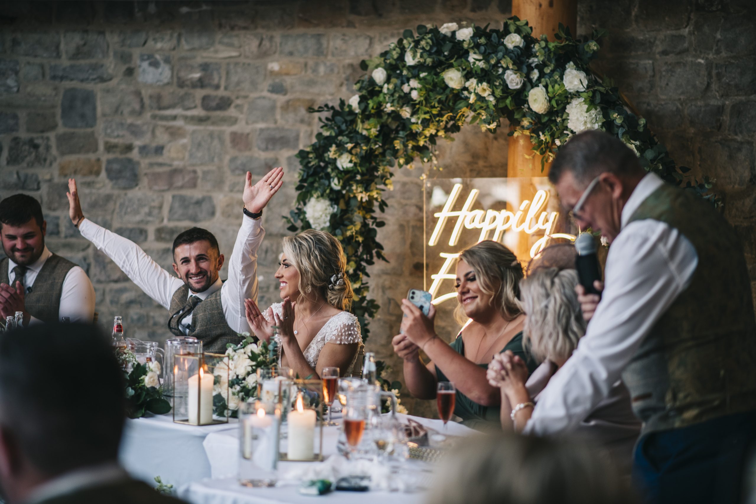 Bridge and groom's head table with groom celebrating with hands in the air whilst father gives speach