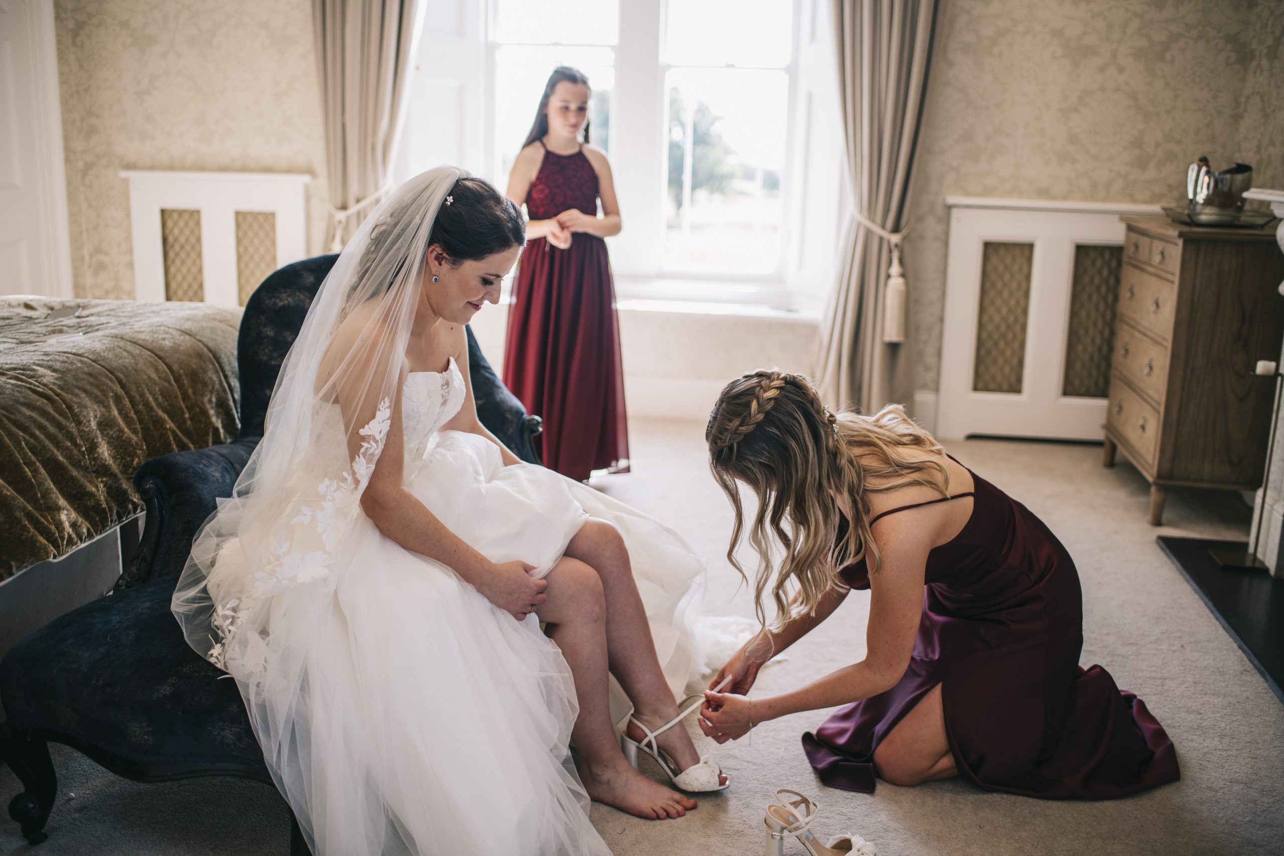 Bride sat on chat with bridesmaid on hands and knees putting her shoes on for her. 